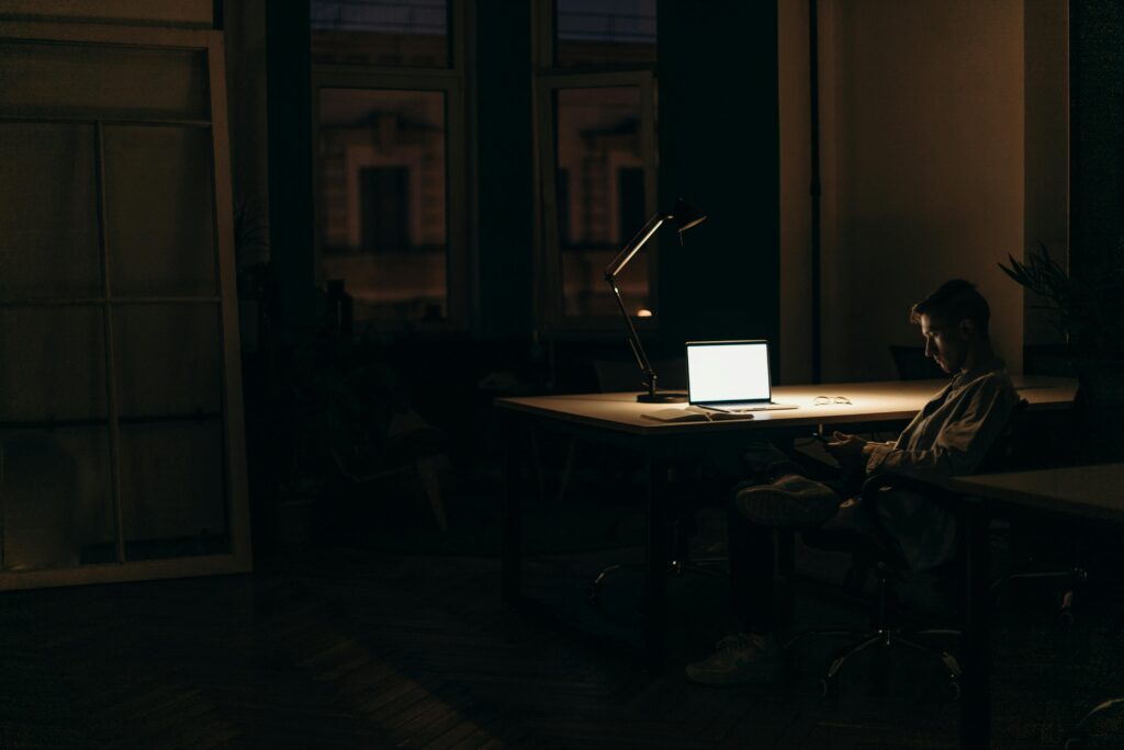 A man working late at night in a dimly lit office, with a bright laptop screen.