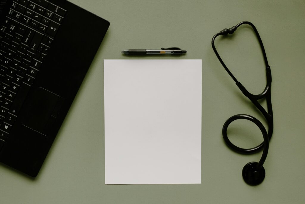 Medical workspace featuring a blank paper, stethoscope, and laptop from above.